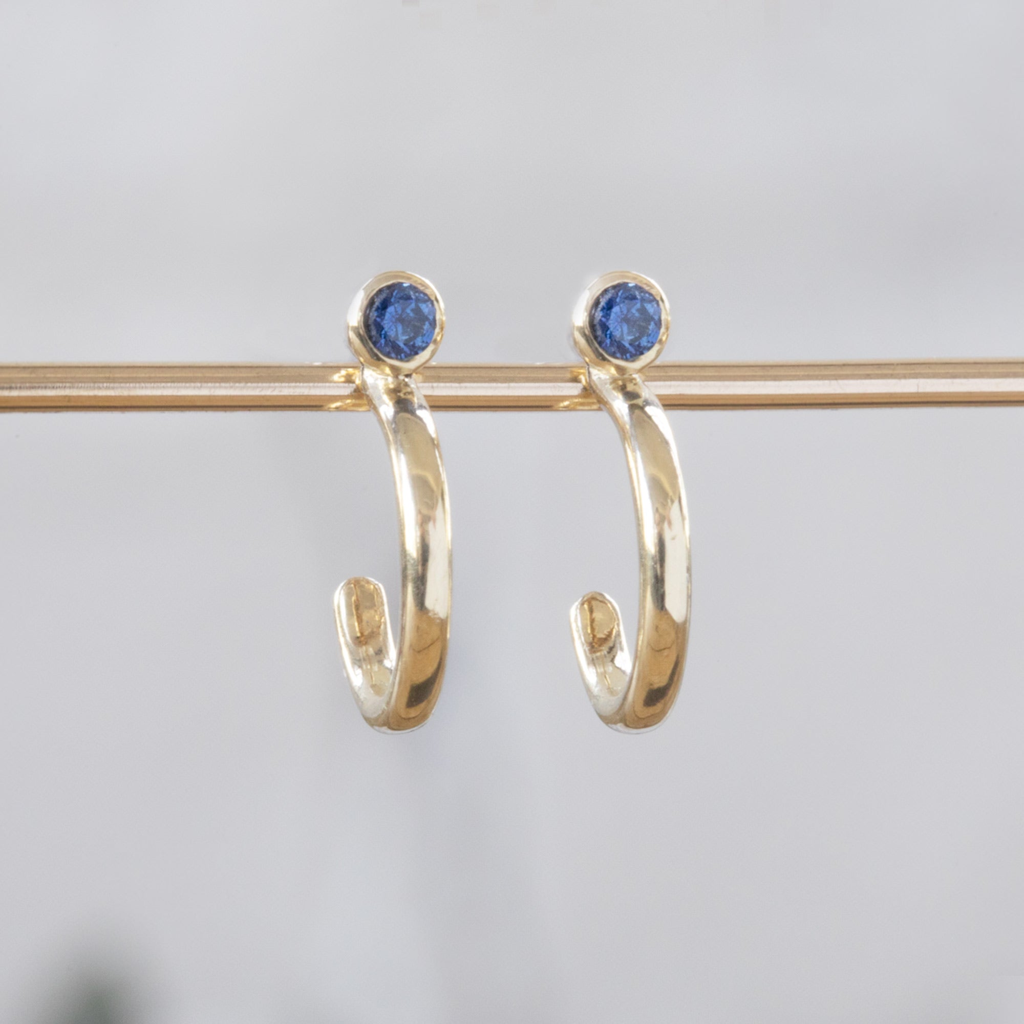 Custom Blue Sapphire Birthstone Hoop Earrings | 14K Yellow Gold displayed on bar with blurred white background.