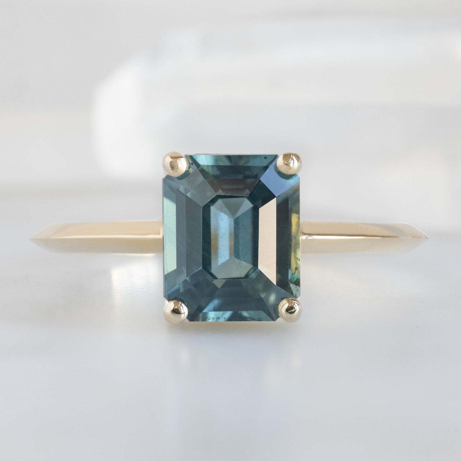 The Bryn Ring with a 1.78ct Emerald Cut Sapphire in 14K Yellow Gold on White Marble
