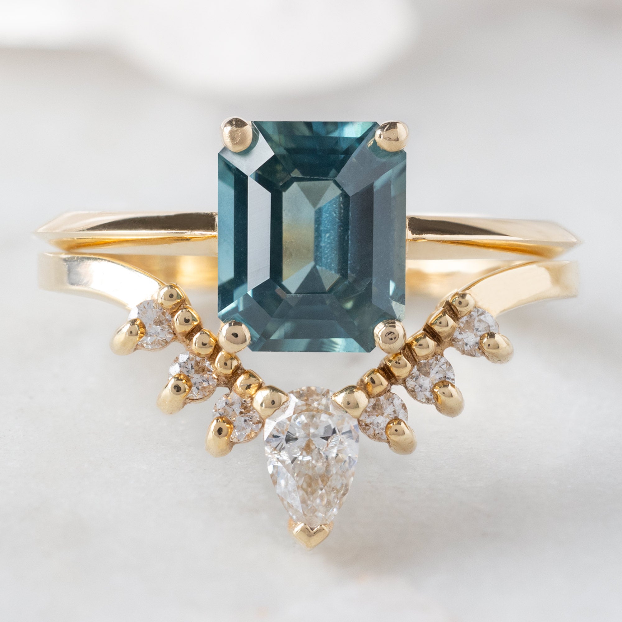 The Bryn Ring with a 1.78ct Emerald Cut Sapphire in 14K Yellow Gold Stacked with the White Diamond Sunburst Stacking Band on White Marble