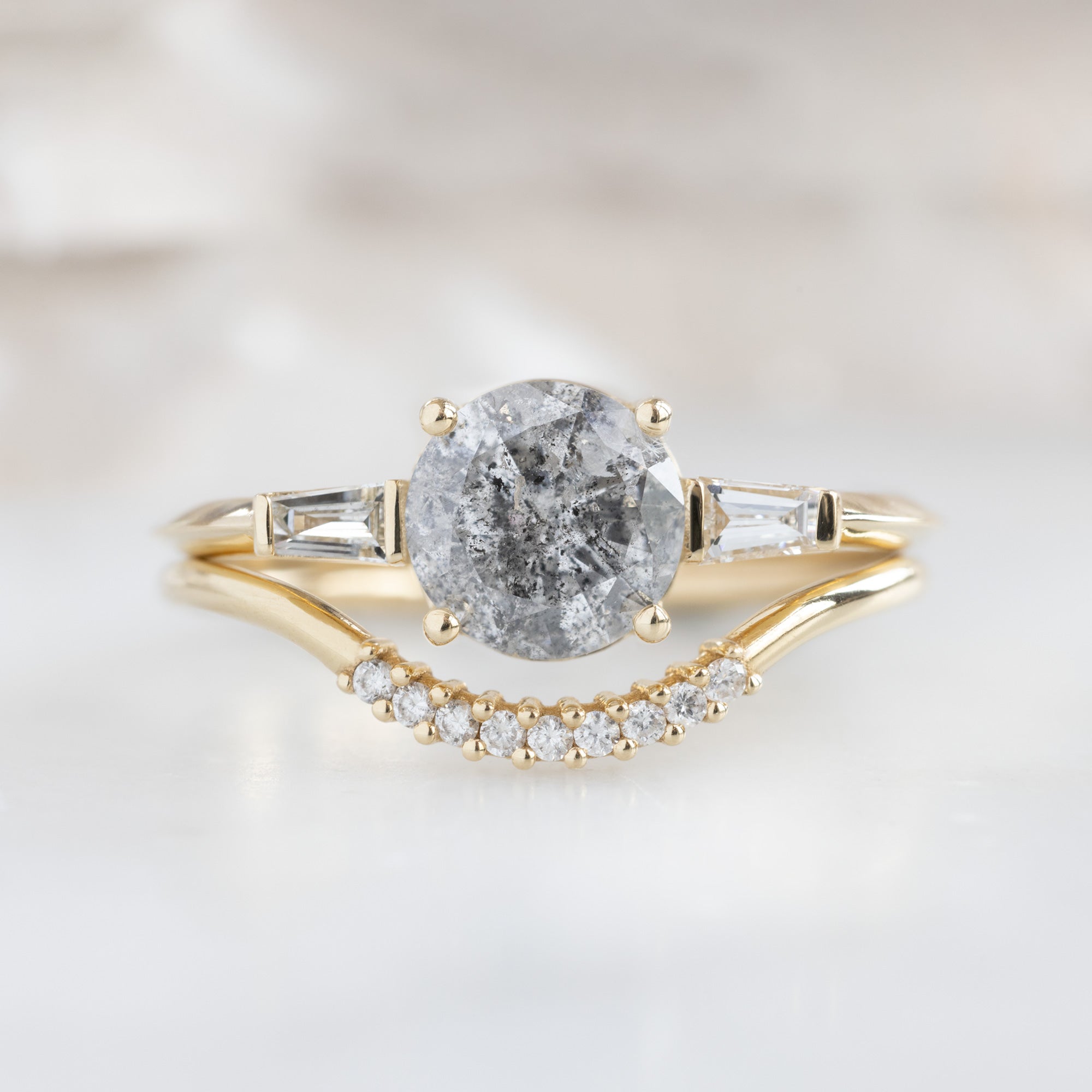 The Ash Ring | 1.22 carat Round Salt and Pepper Diamond in 14K Yellow Gold with white blurred background. Displayed upon the white diamond pavé arc stacking ring.