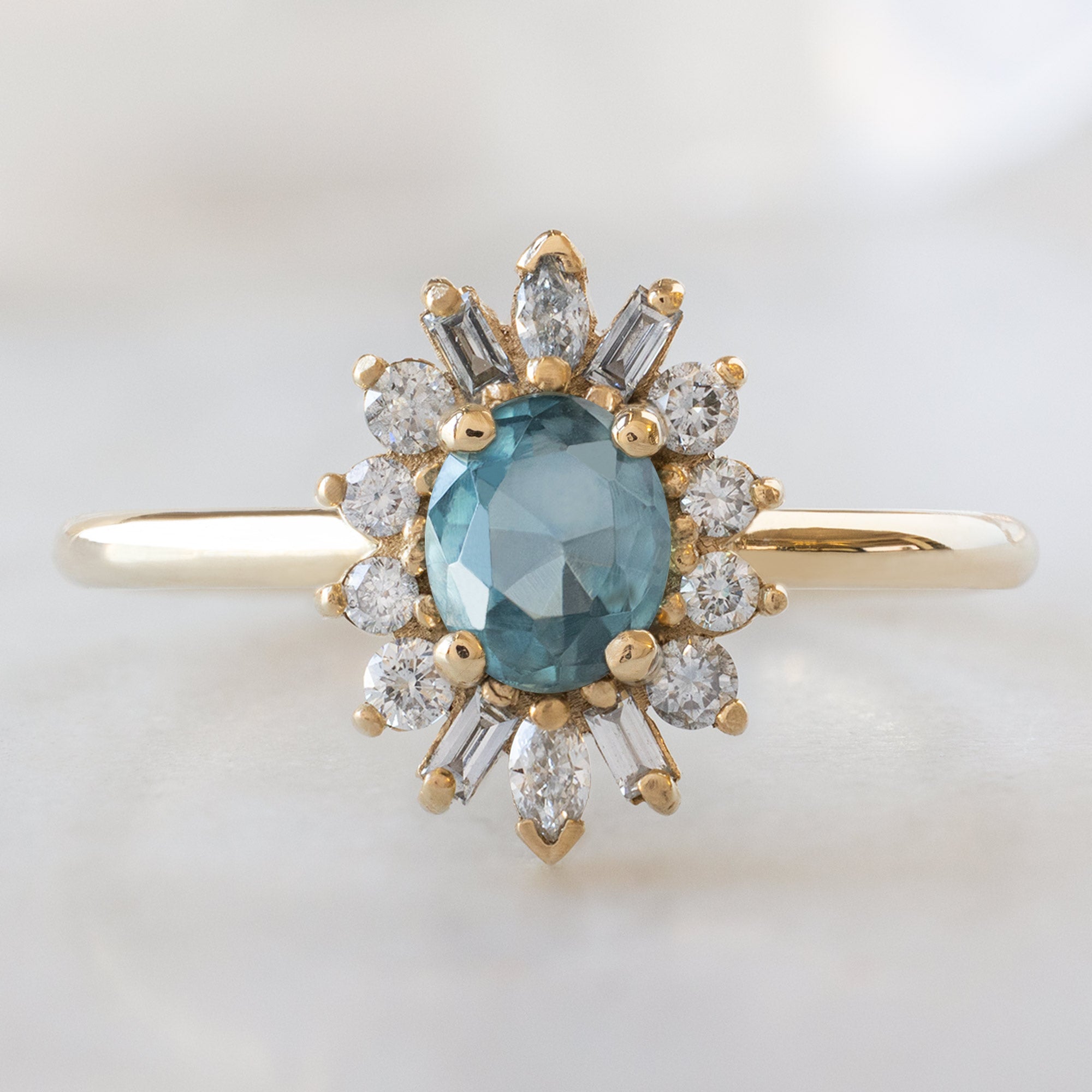 The Camellia Ring with a 0.81ct Oval Blue Montana Sapphire in 14K Yellow Gold on White Marble