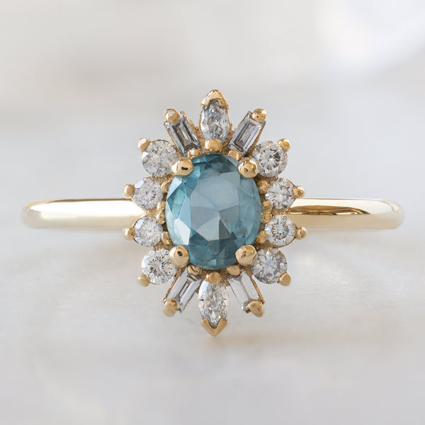 The Camellia Ring with an Oval Cut Blue Sapphire in 14K Yellow Gold on White Marble