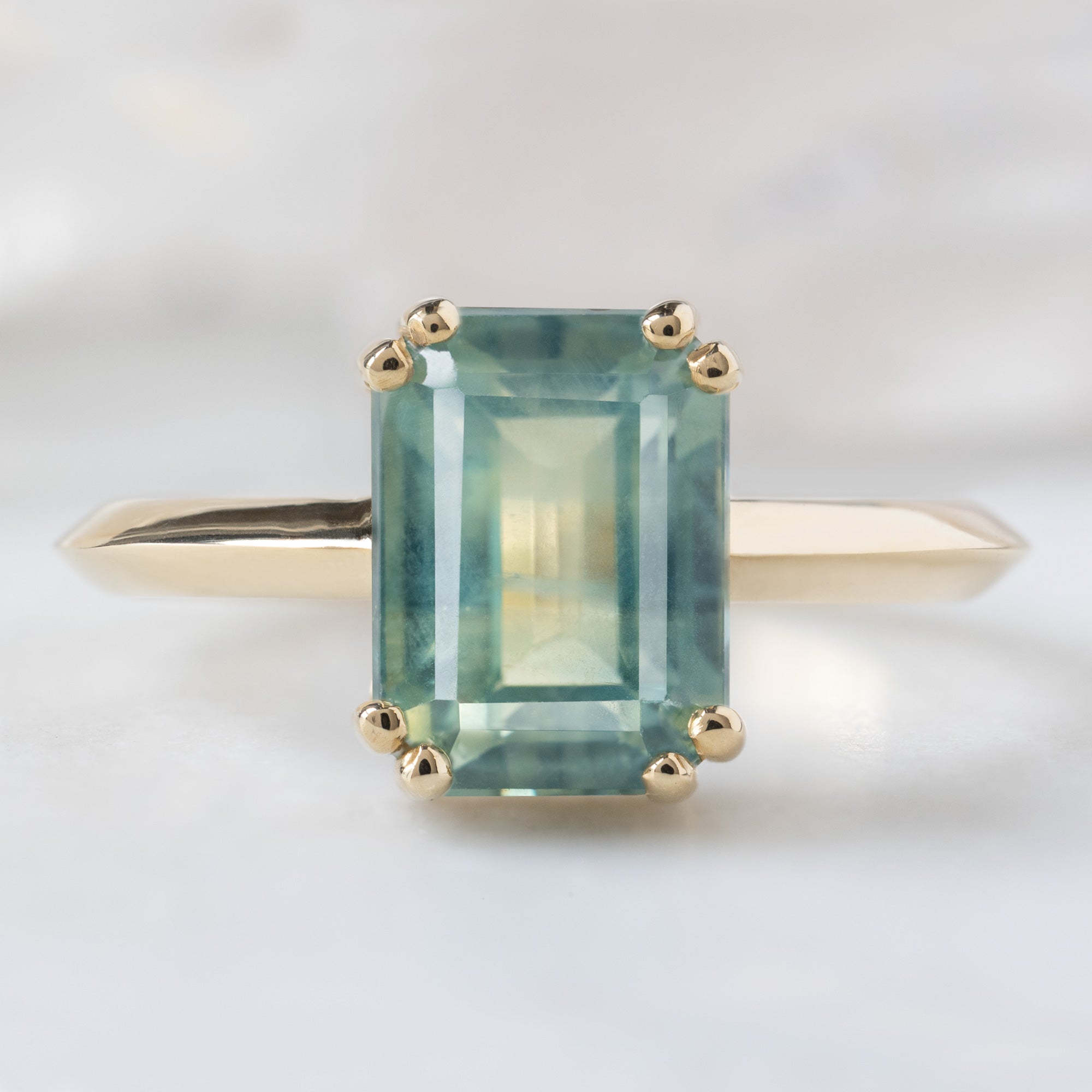 The Bryn Ring with a 2.97ct Emerald Cut Bi-Color Sapphire in 14K Yellow Gold on White Marble Tile