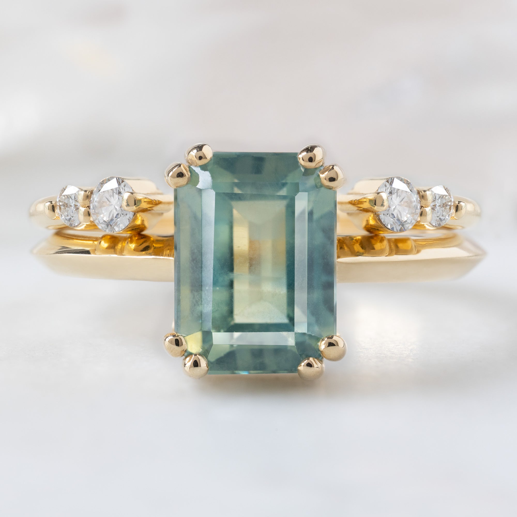 The Bryn Ring with a 2.97ct Emerald Cut Bi-Color Sapphire in 14K Yellow Gold Stacked with the Luxe Open Cuff Round White Diamond Stacking Ring on White Marble Tile