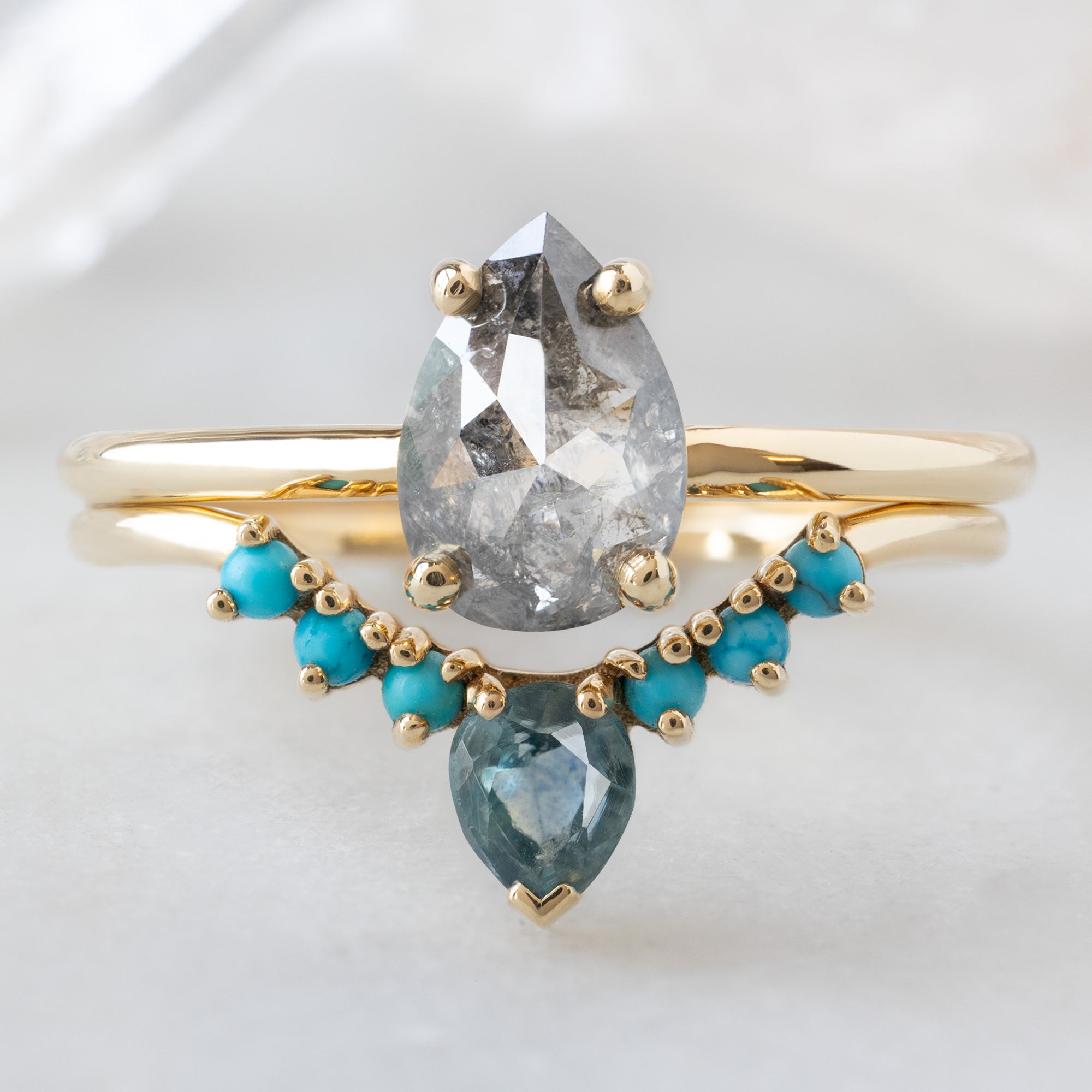 The Bryn Ring with a 1.01ct Pear Salt and Pepper Diamond in 14K Yellow Gold  Stacked with the Montana Sapphire and Turquoise Sunburst Stacking Band on White Marble Surface
