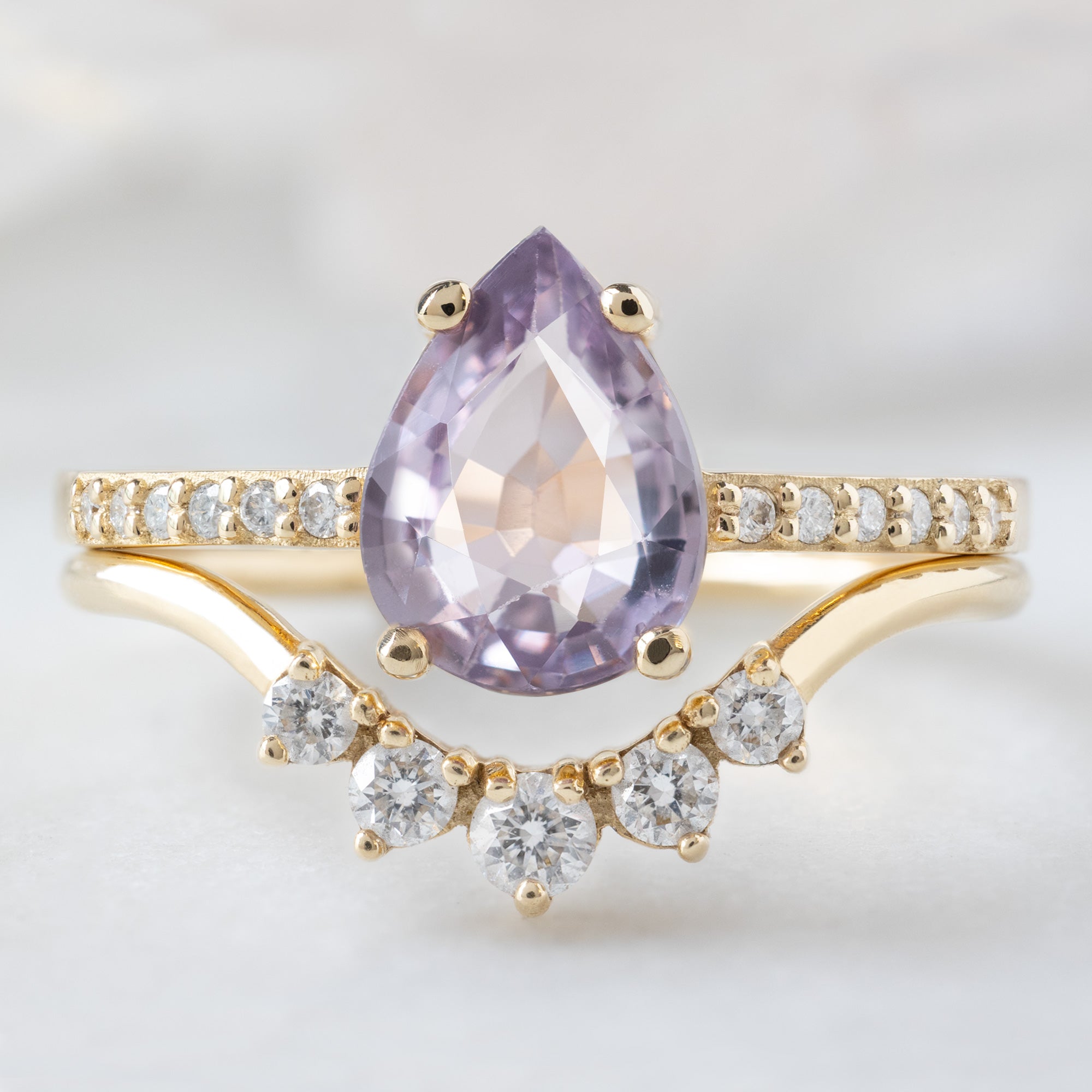 The Willow Ring with a 1.38ct Pear Violet Sapphire in 14K Yellow Gold Stacked with the Round White Diamond Sunburst Stacking Band on White Marble