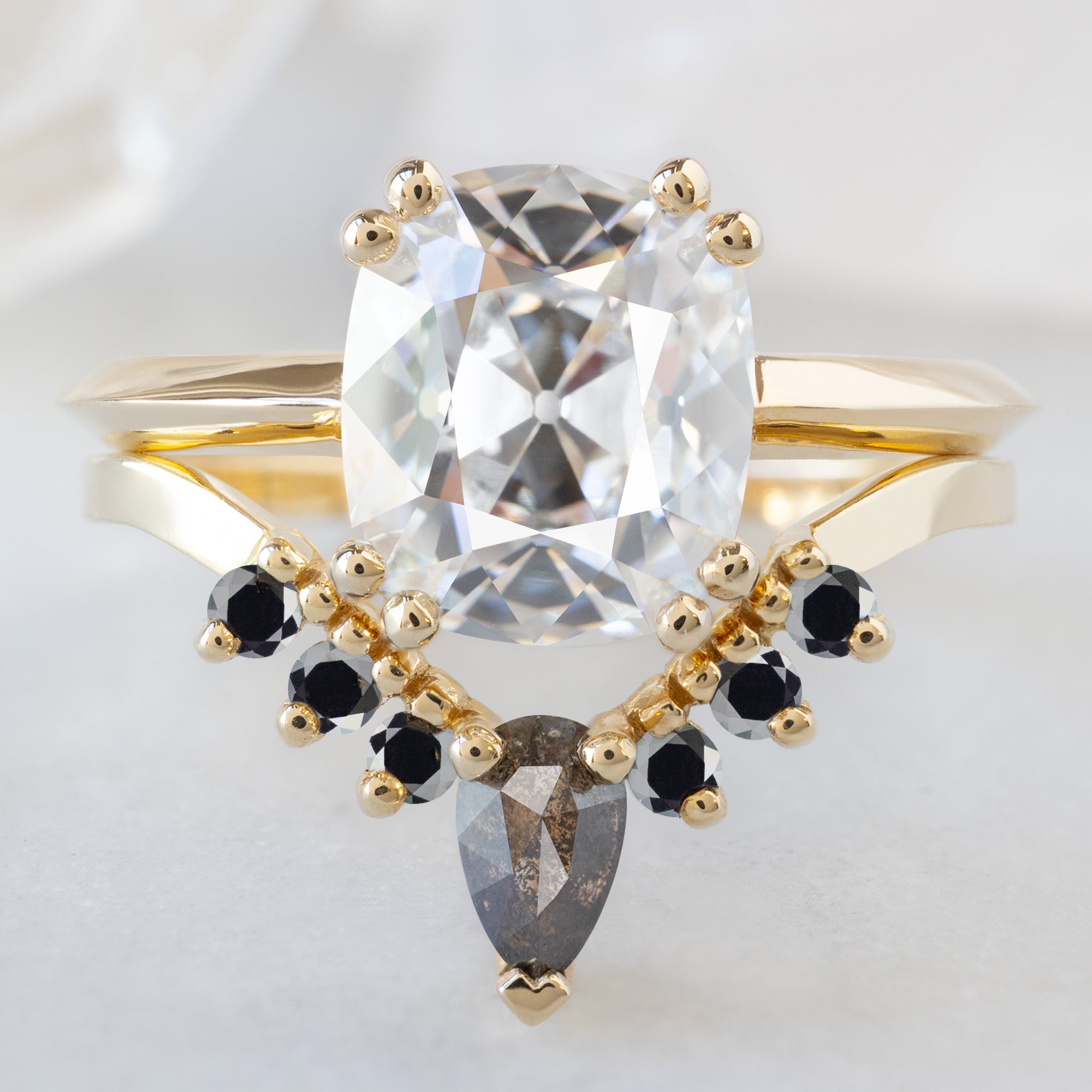 The Bryn Ring with a 2.32ct Old Mine Cut White Diamond in 14K Yellow Gold Stacked with the Black Diamond Sunburst Stacking Band on White Marble Surface