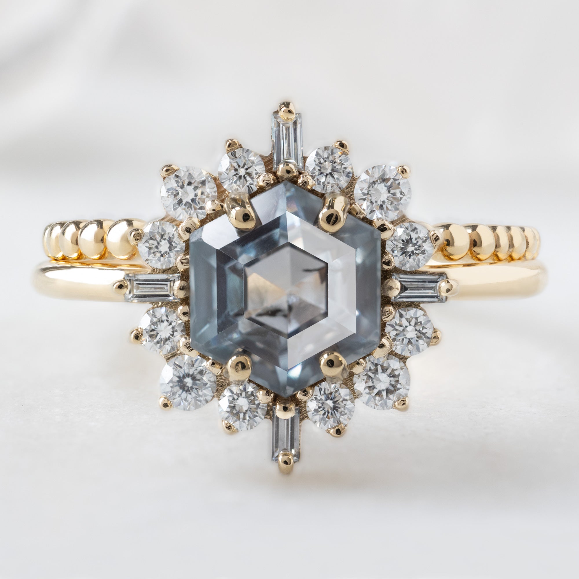 The Compass Ring with a 1.67ct Hexagon Grey Sapphire in 14K Yellow Gold Stacked with the Beaded Stacking Band on White Marble Surface