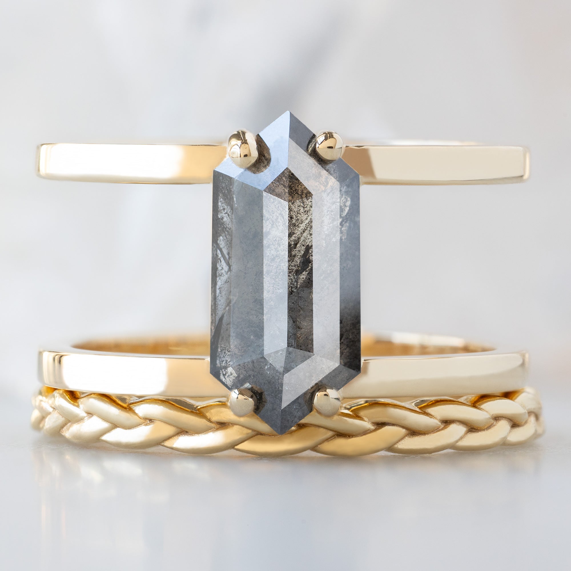The Poppy Ring with a 1.57ct Hexagon Salt and Pepper Diamond in 14K Yellow Gold Stacked with Braided Stacking Band on White Marble Surface