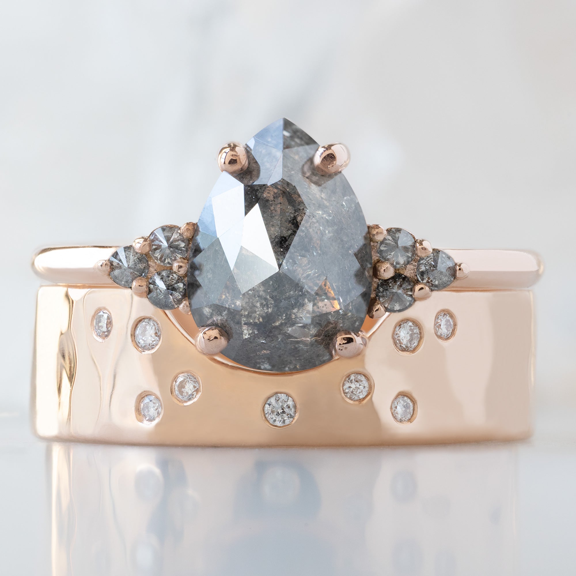 The Ivy Ring with a 1.43ct Pear Salt and Pepper Diamond in 14K Rose Gold Stacked with the Constellation Arc Stacking Band on White Marble Surface