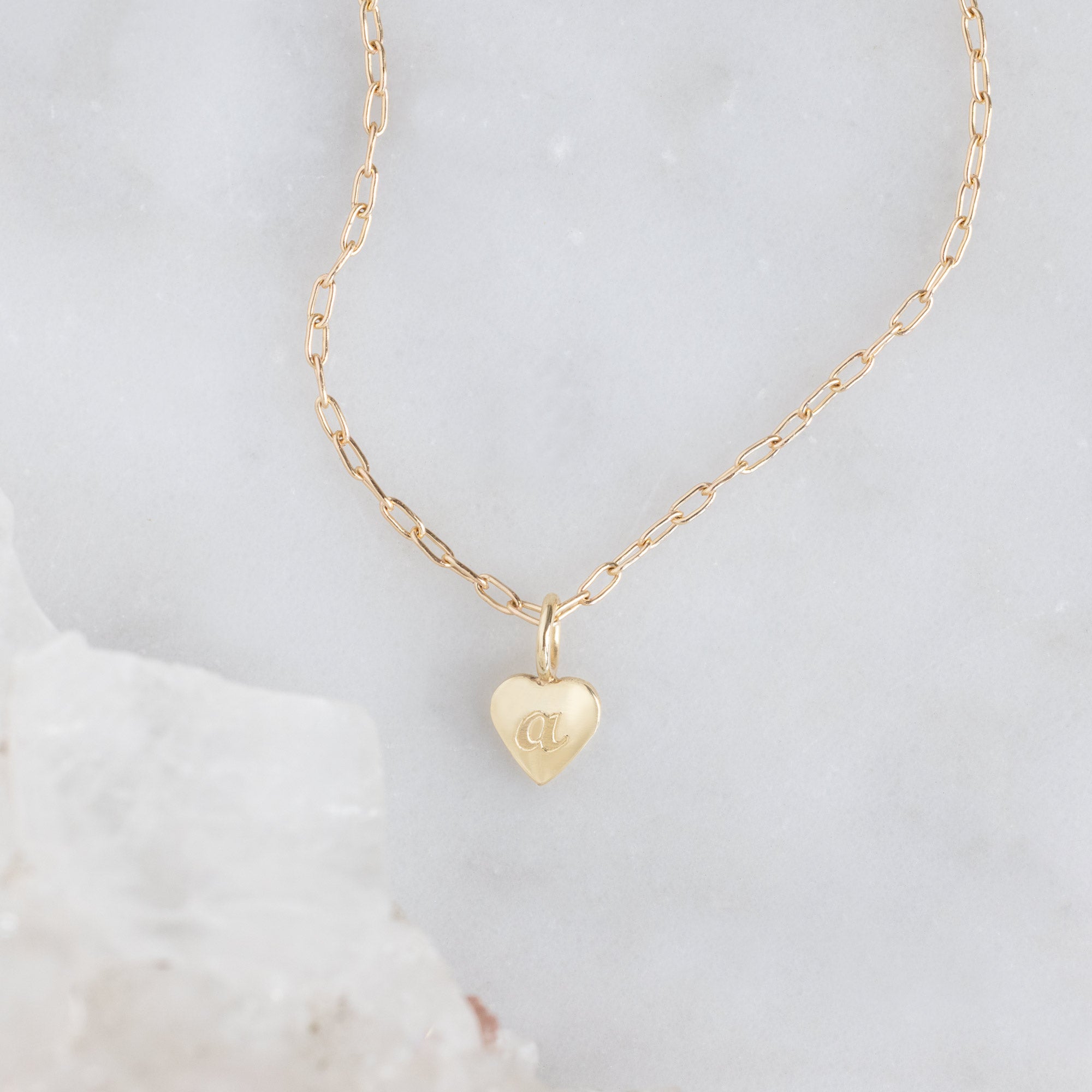 The Heart Pendant Necklace with an engraved 'A' in Yellow Gold on White Marble Surface