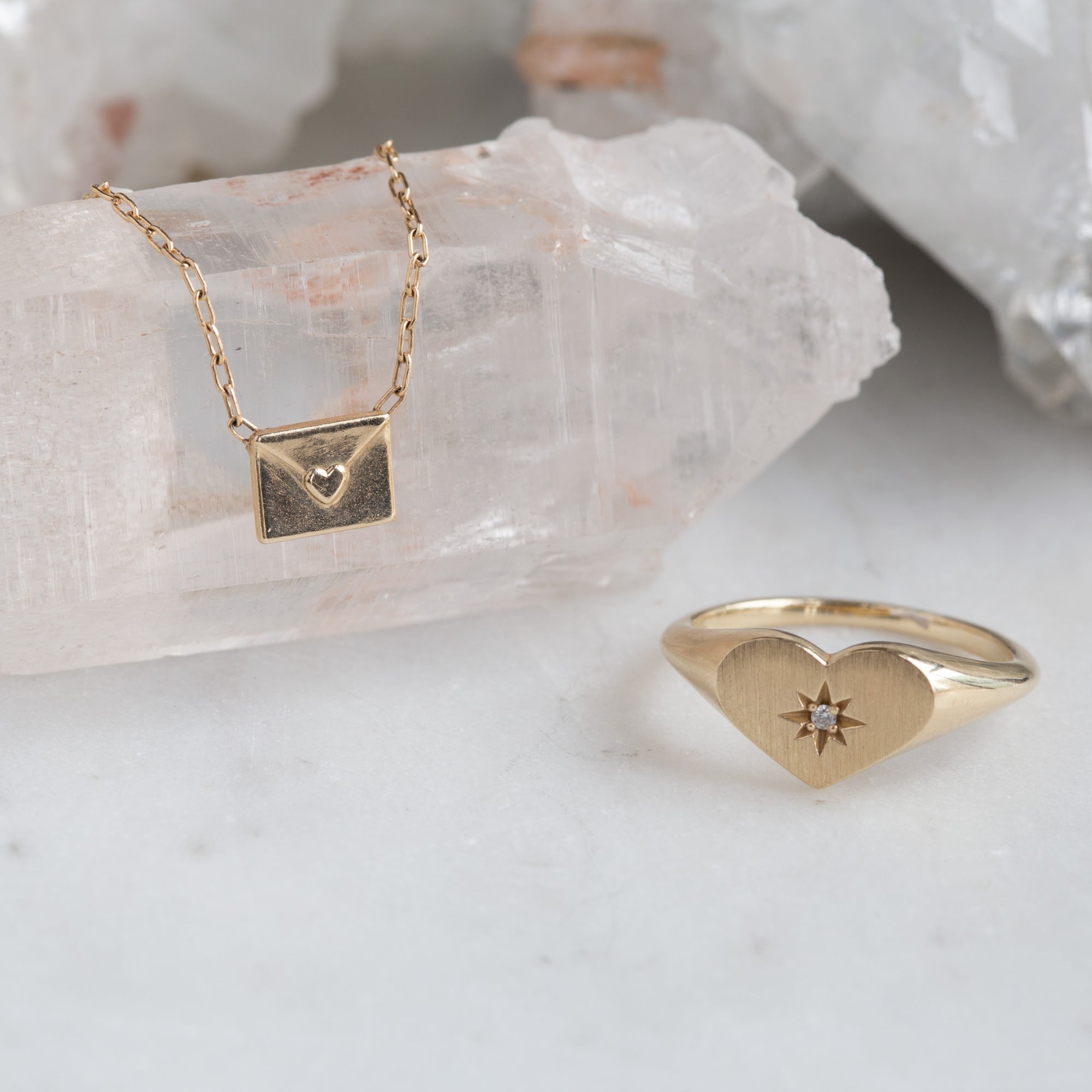 yellow gold necklace featuring a loveletter shaped pendant hanging on white crystal next to heart shaped signet ring