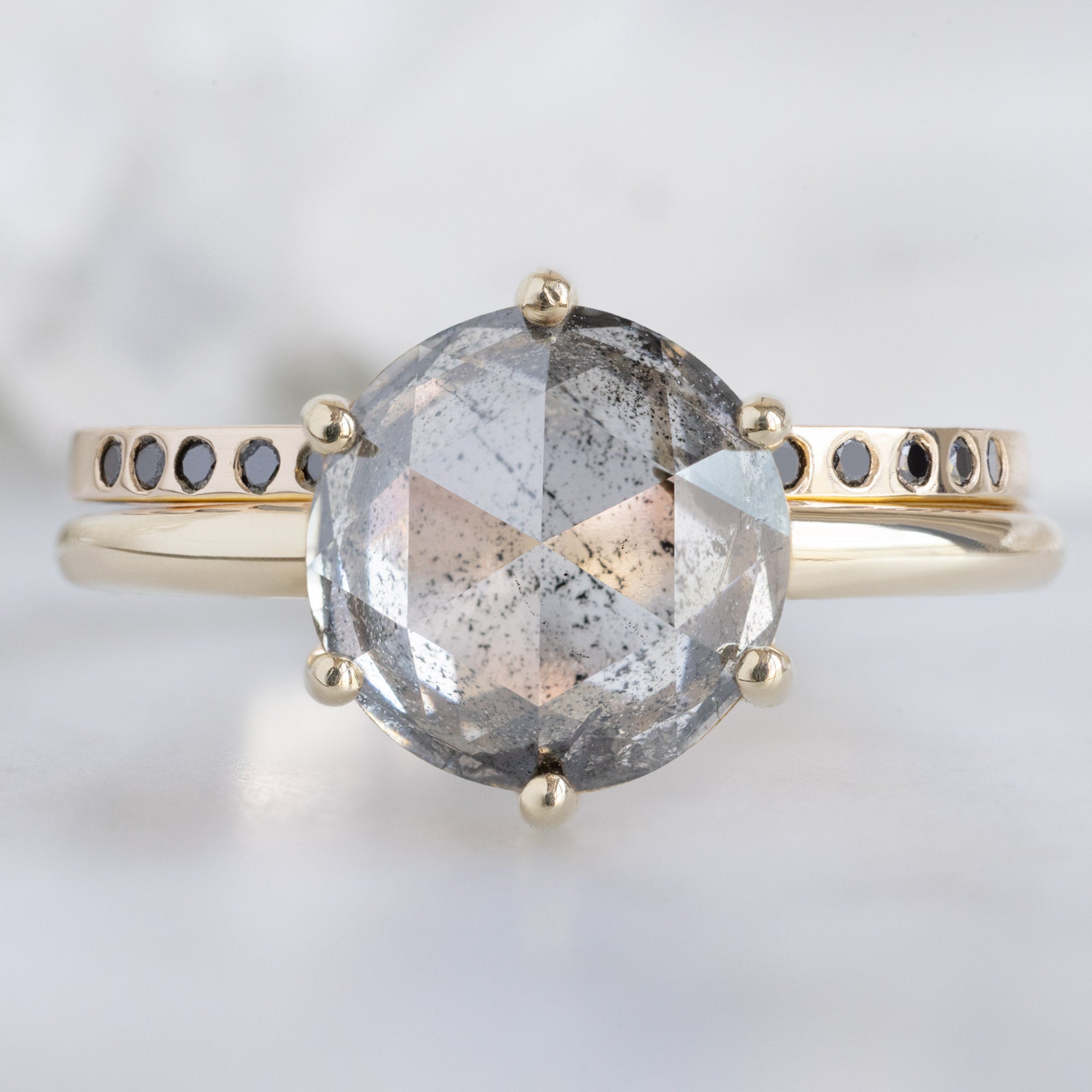 Open Cuff Flush Set Black Diamond Pavé Stacking Band in 14K Rose Gold Stacked with Round Salt and Pepper Diamond Solitaire Engagement Ring on White Marble Surface