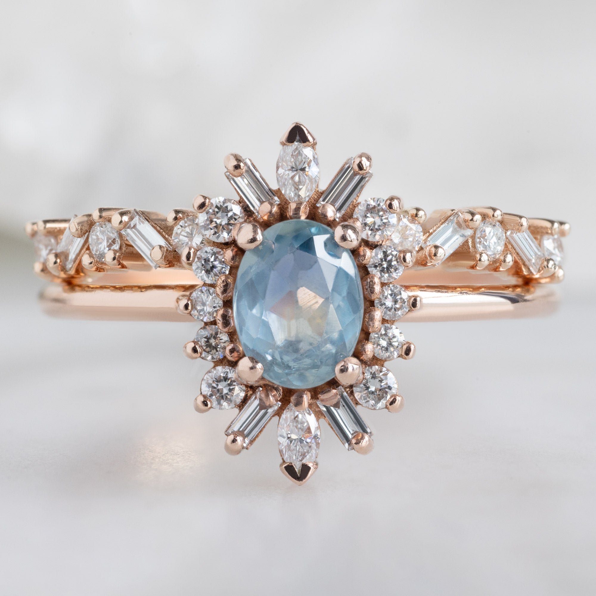 The Baguette Confetti Stacking Ring In 14k Rose Gold Gold Stacked with Blue Sapphire Oval Engagement Ring on White Marble Surface