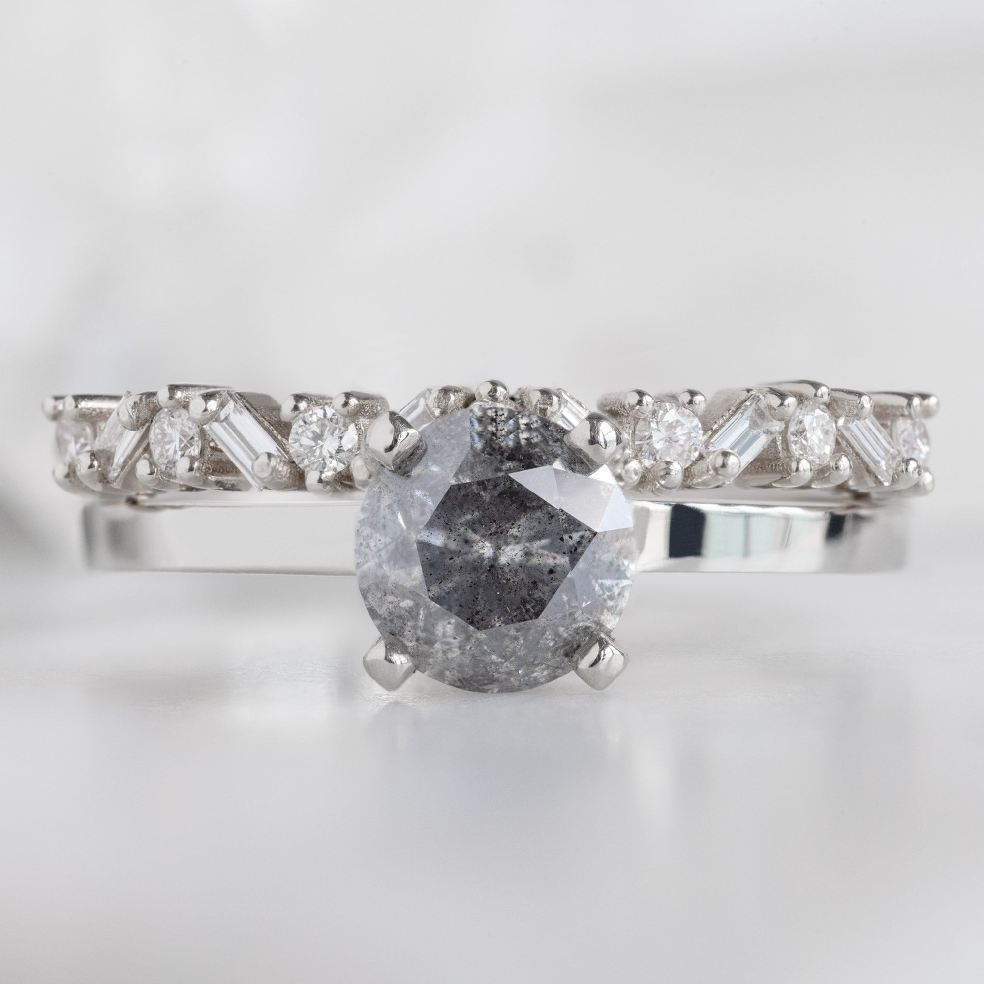 The Baguette Confetti Stacking Ring In 14k White Gold Stacked with Round Salt and Pepper Diamond Engagement Ring on White Marble Surface