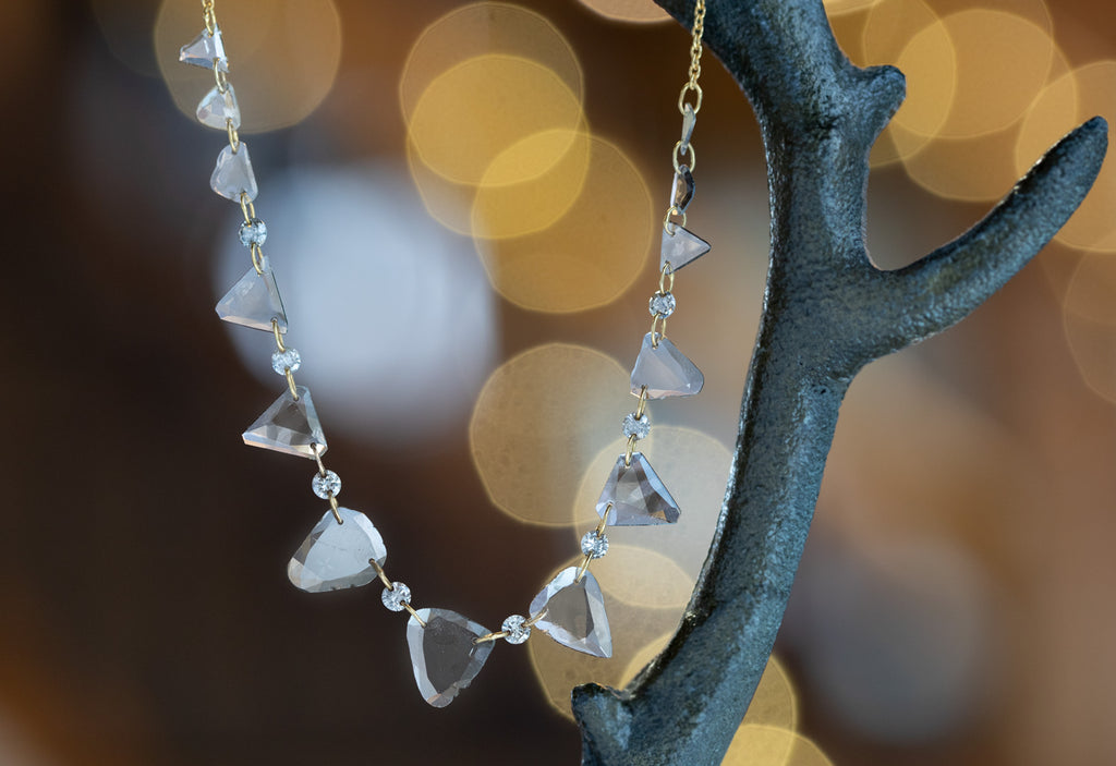 Yellow Gold Diamond Slice Pennant Necklace hanging on steel earring tree with blurred white string lights in background