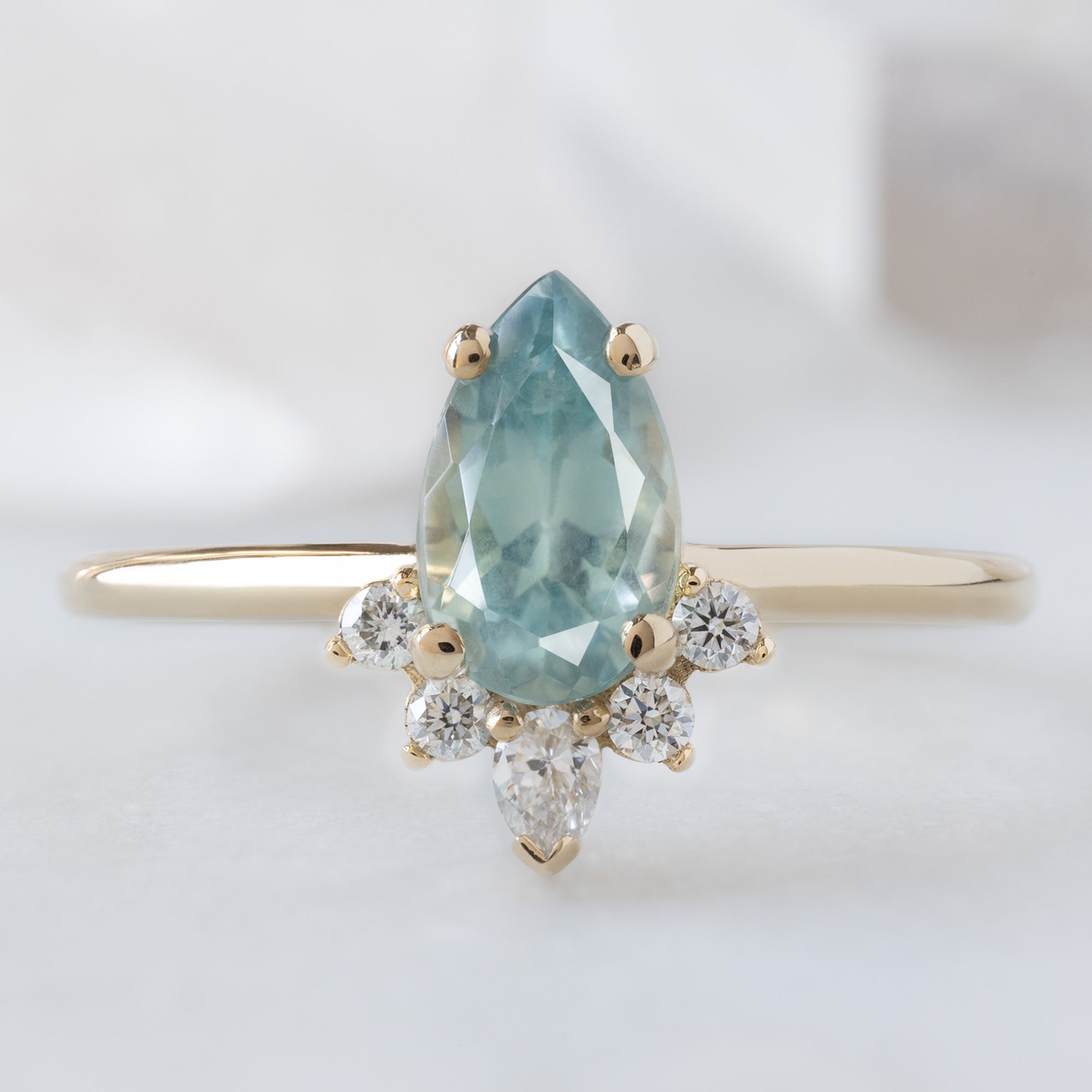 The Aster Ring with a 1.18ct Pear Blue Sapphire in 14K Yellow Gold on White Marble