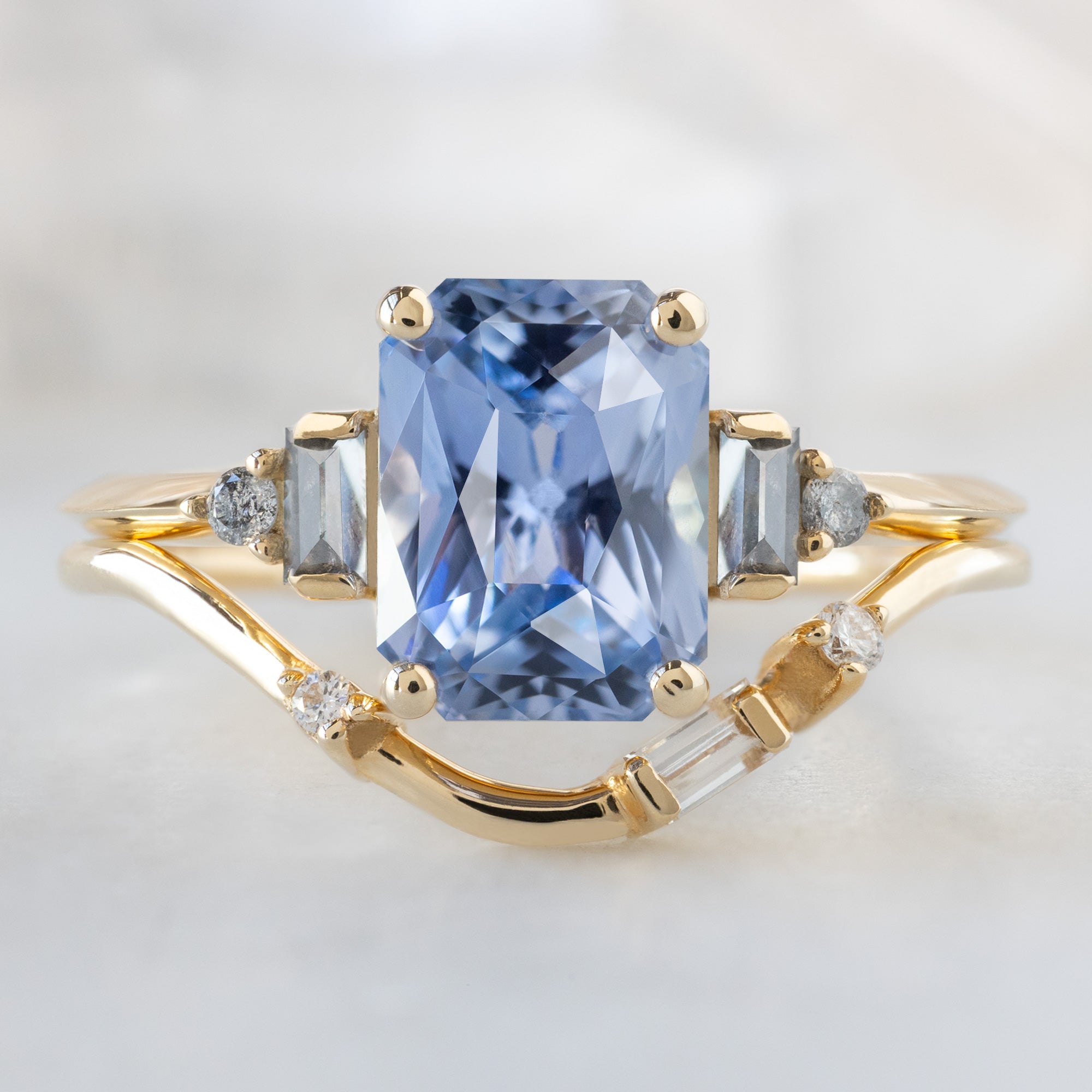 The Primrose Ring with a 2.36ct Emerald Cut Sapphire in 14K Yellow Gold Stacked with the Asymmetrical Baguette Diamond Stacking Band on White Marble Surface