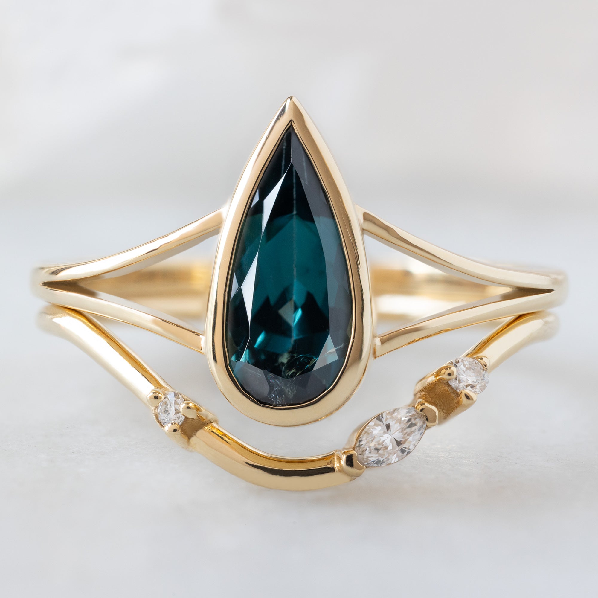 The Magnolia Ring with a 1.48ct Pear Blue Sapphire in 14K Yellow Gold Stacked with the Asymmetrical Arc Marquise Stacking Band on White Marble Surface