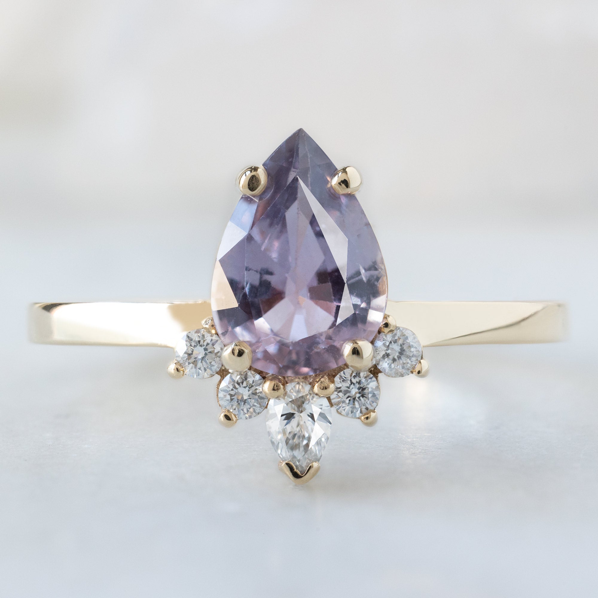 The Aster Ring with a 1.06ct Pear Purple Sapphire in 14K Yellow Gold on White Marble Surface