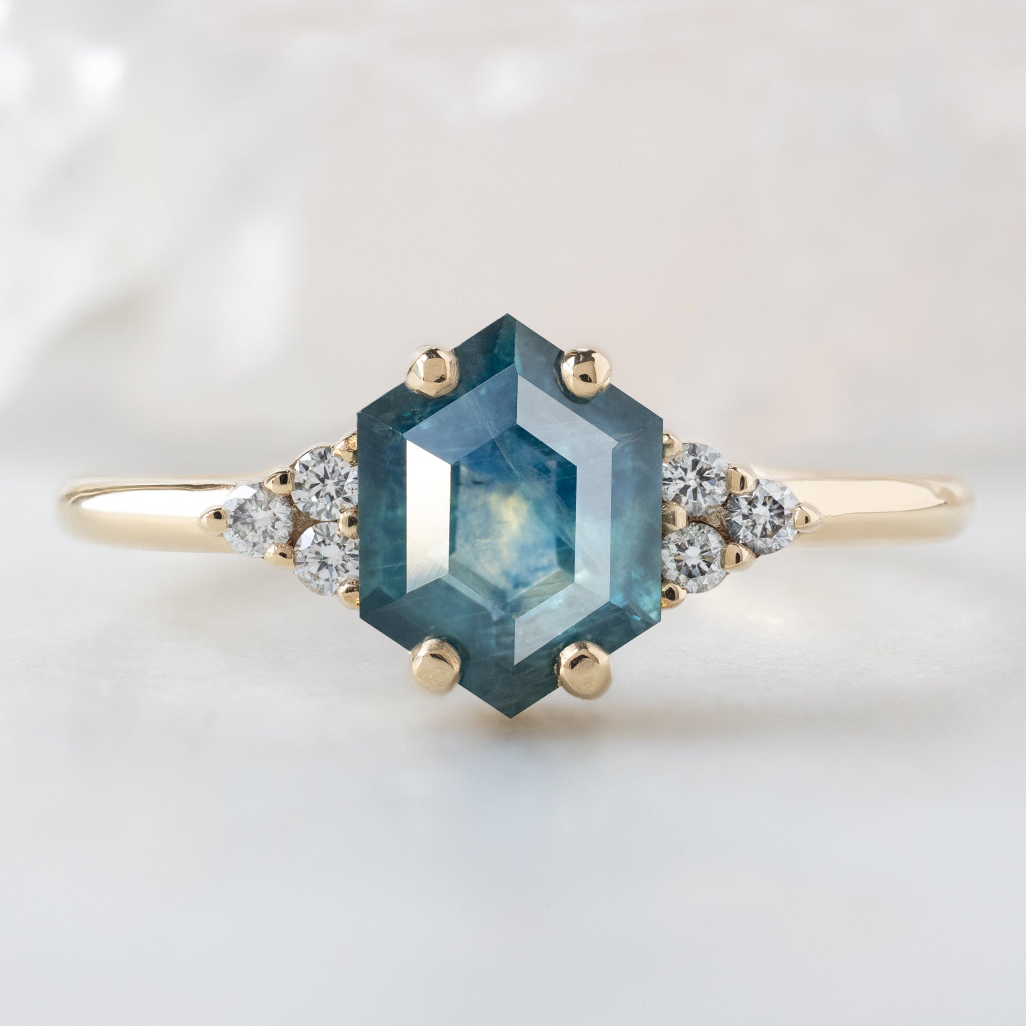 The Ivy Ring with a 1.22ct Hexagon Blue Sapphire in 14K Yellow Gold on White Marble Surface