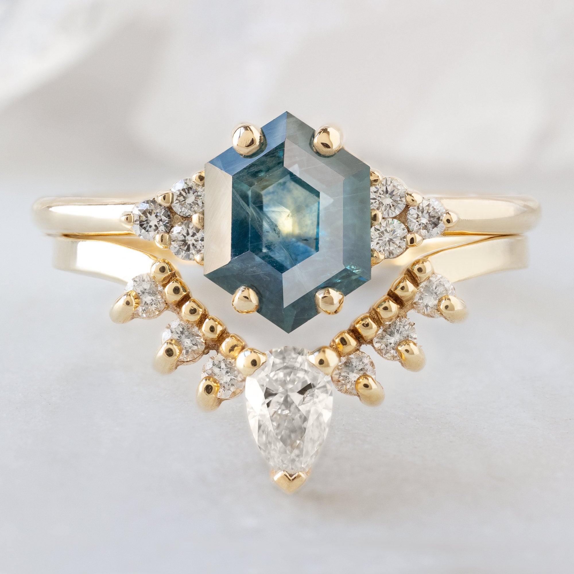 The Ivy Ring with a 1.22ct Hexagon Blue Sapphire in 14K Yellow Gold Stacked with the White Diamond Sunburst Stacking Band on White Marble Surface