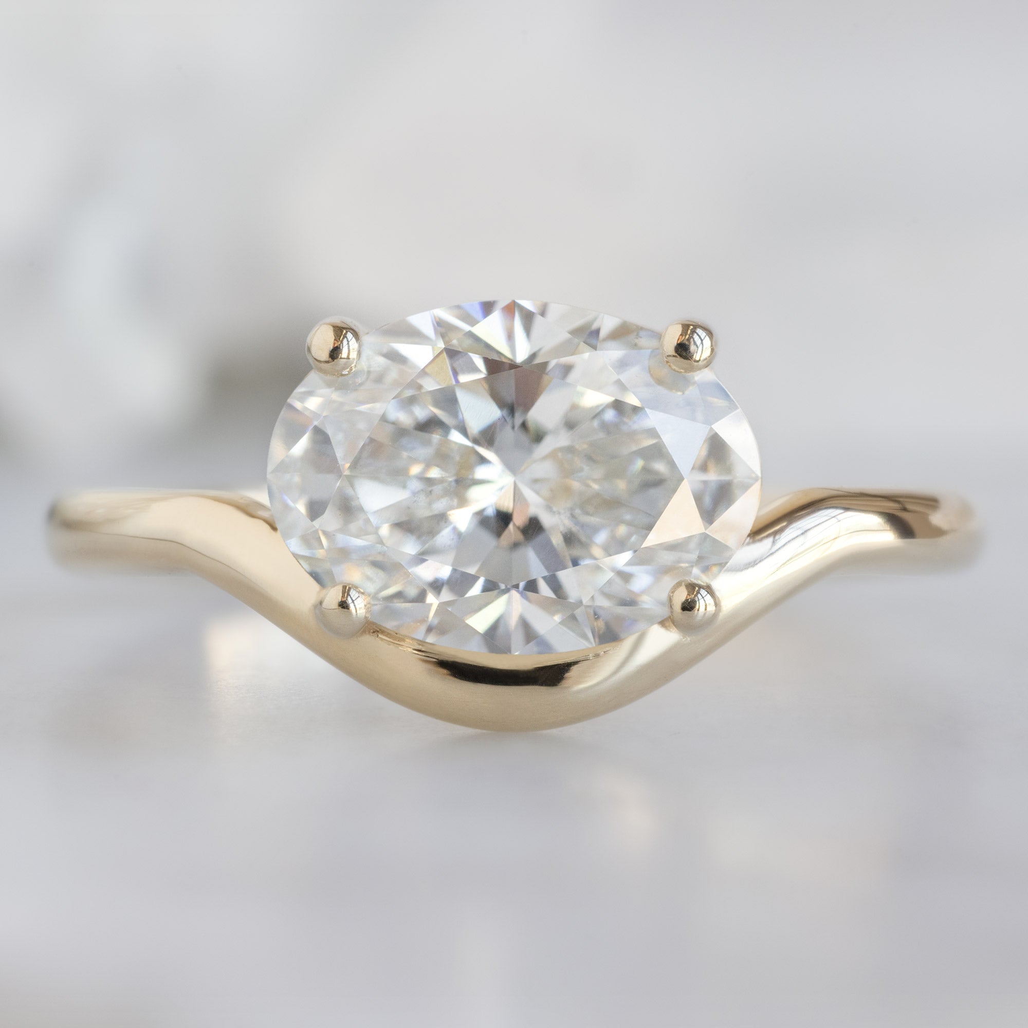 The Wave Ring with a 1.59ct Oval Lab Grown White Diamond in 14K Yellow Gold on White Marble Surface