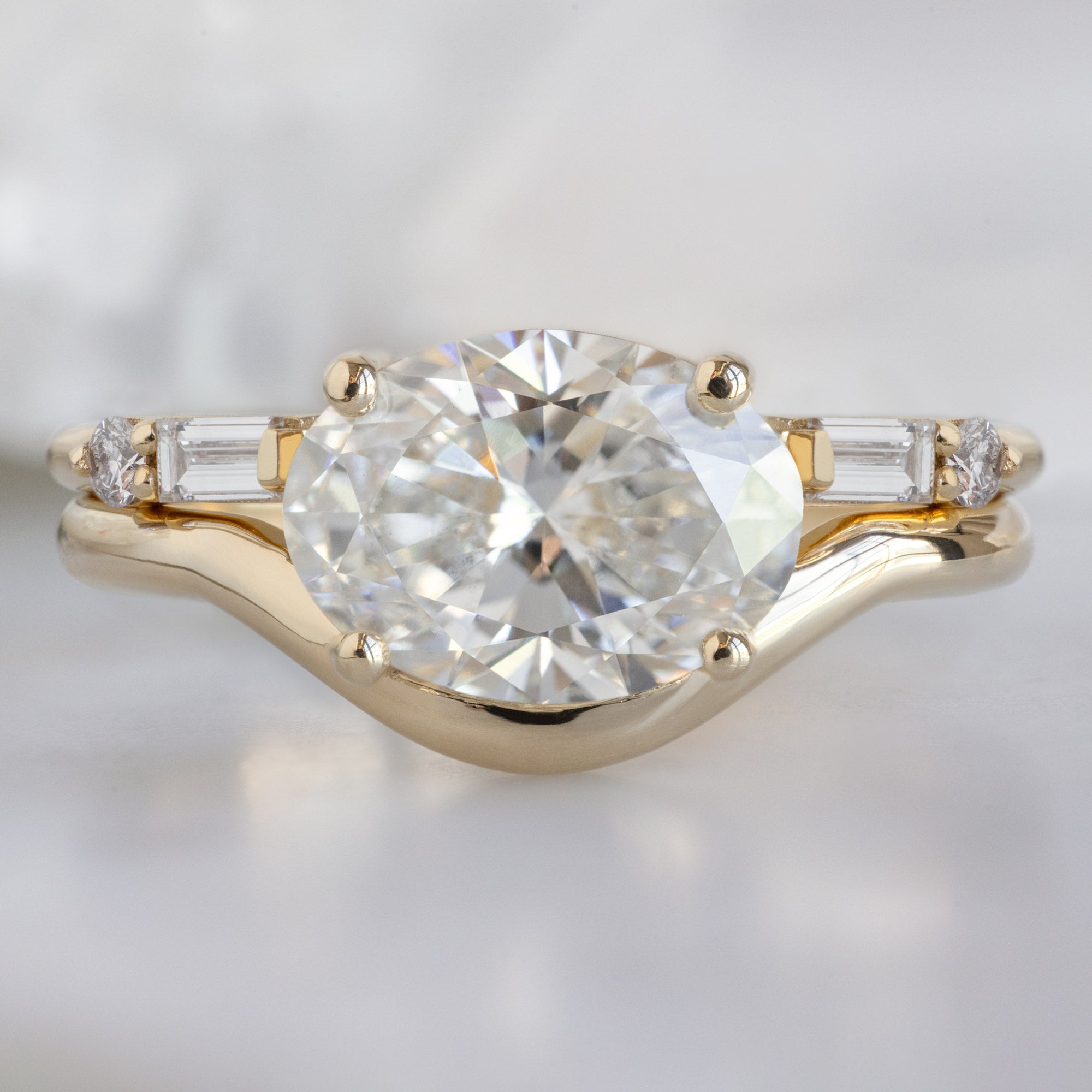 The Wave Ring with a 1.59ct Oval Lab Grown White Diamond in 14K Yellow Gold Stacked with the Open Cuff Luxe White DiamondBaguette Stacking Band on White Marble Surface