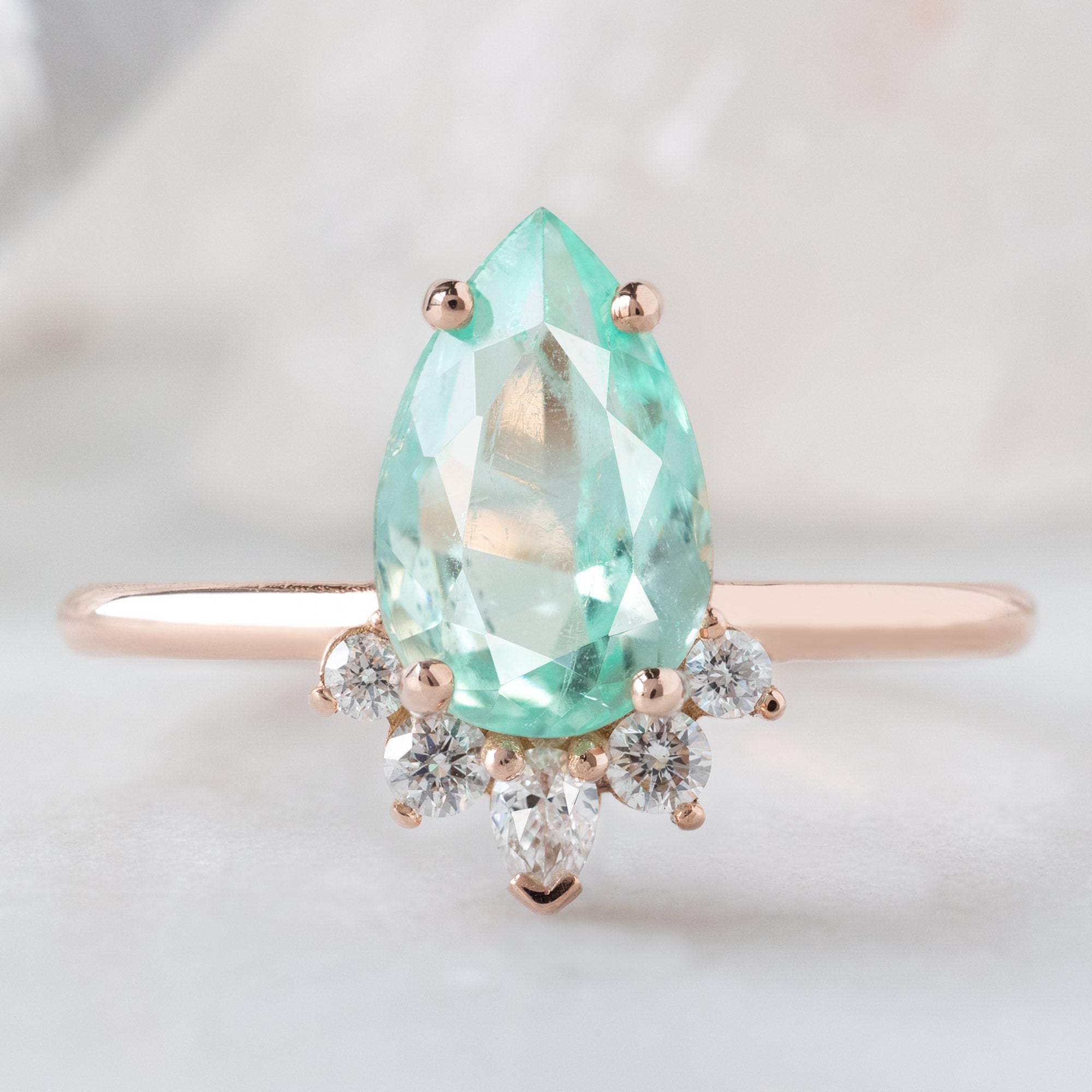 The Aster Ring with a 1.54ct Pear Emerald in 14K Rose Gold on White Marble Surface
