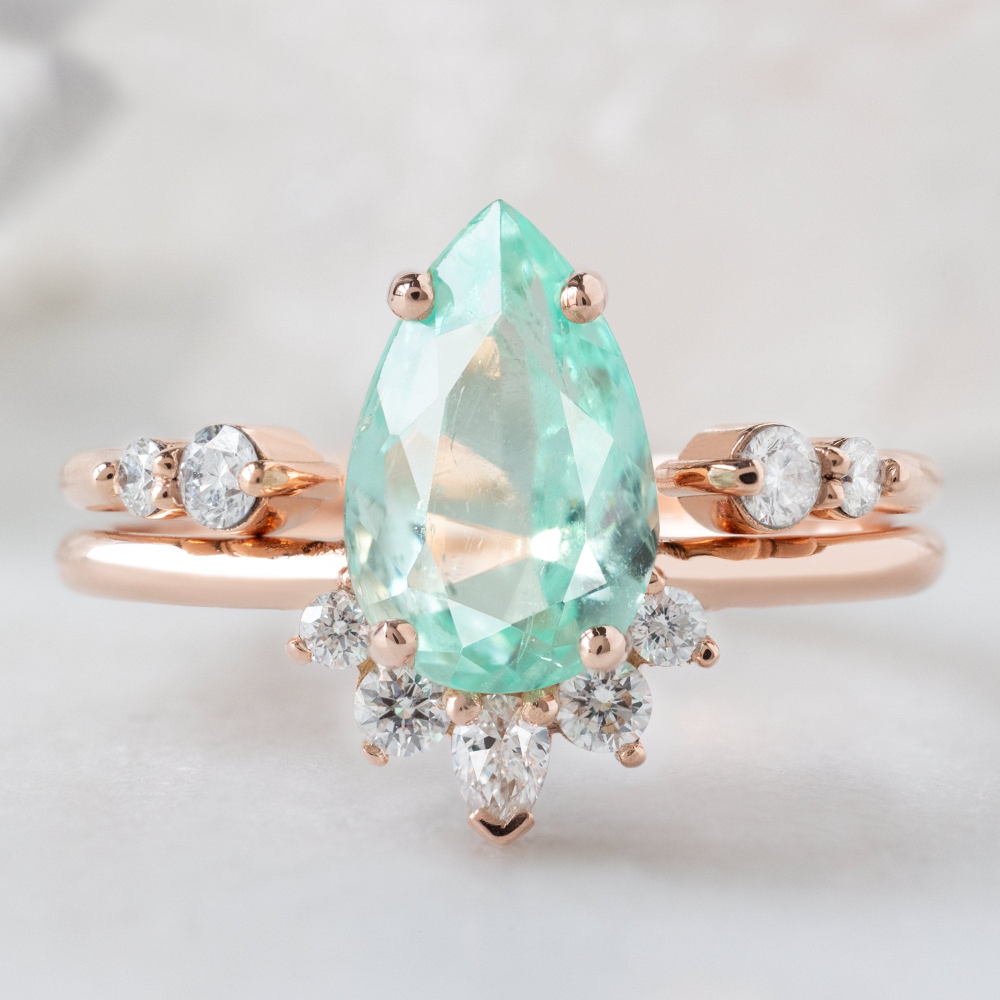 The Aster Ring with a 1.54ct Pear Emerald in 14K Rose Gold Stacked with the Luxe Open Cuff Round White Diamond Stacking Band on White Marble Surface