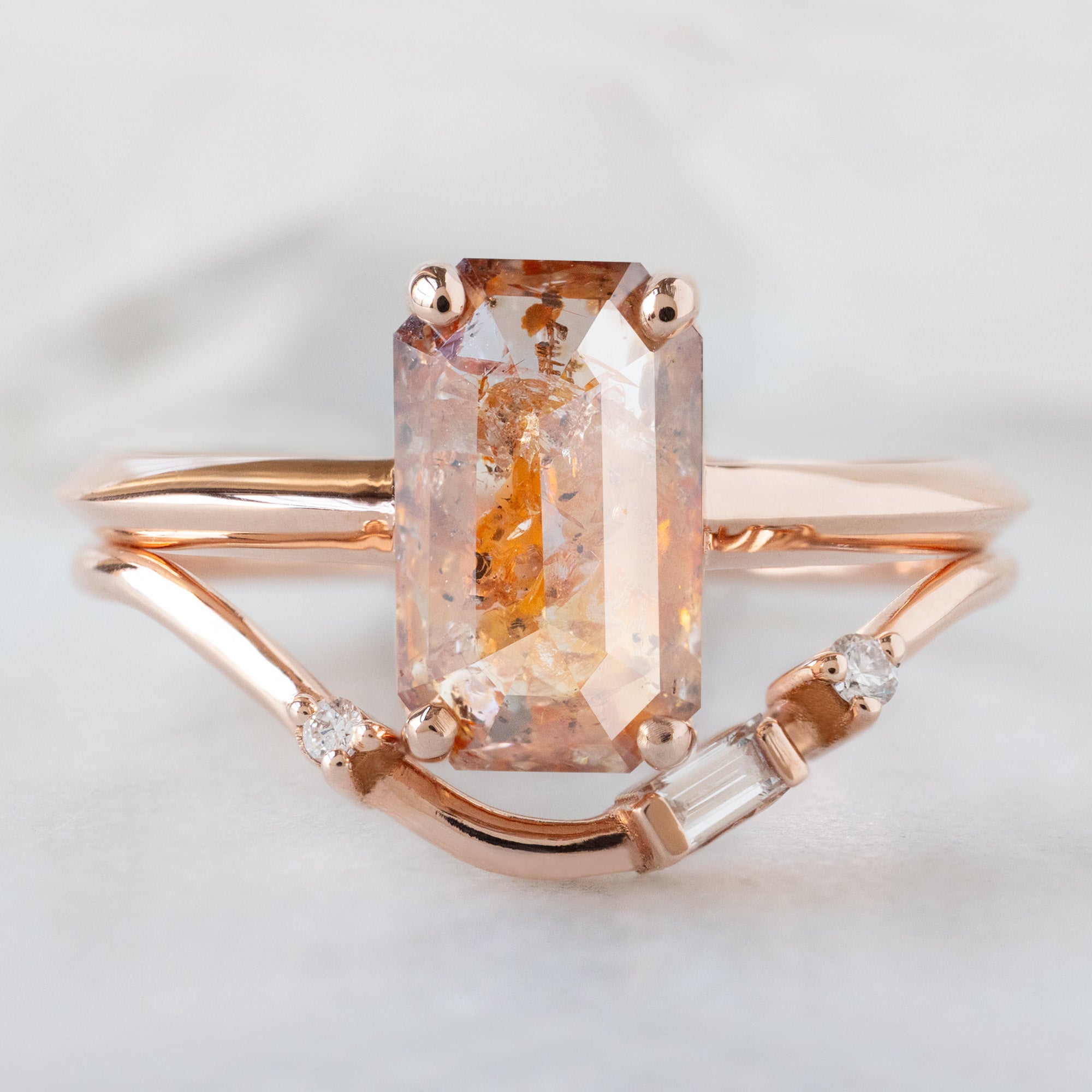 The Bryn Ring with a 2.01ct Emerald Cut Red Diamond in 14K Rose Gold Stacked with the Asymmetrical Baguette Arc Stacking Band on White Marble Surface