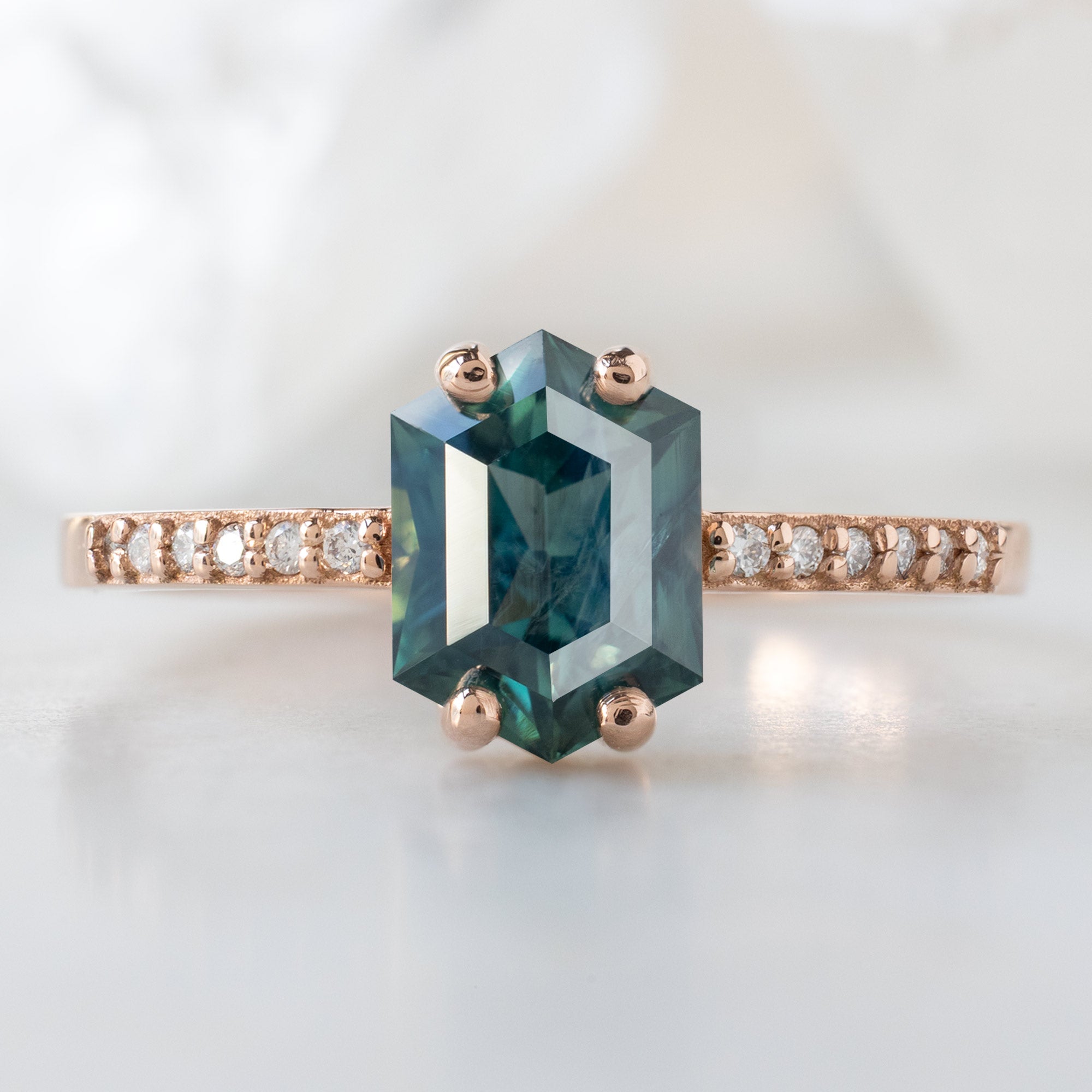 The Willow Ring with a 1.28ct Hexagon Blue Sapphire in 14K Rose Gold on White Marble Surface