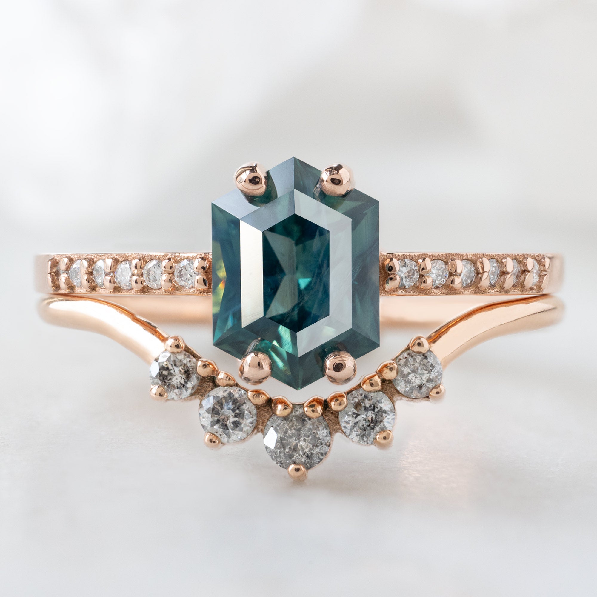 The Willow Ring with a 1.28ct Hexagon Blue Sapphire in 14K Rose Gold on Stacked with the Round Salt and Pepper Diamond Sunburst Stacking Band on White Marble Surface