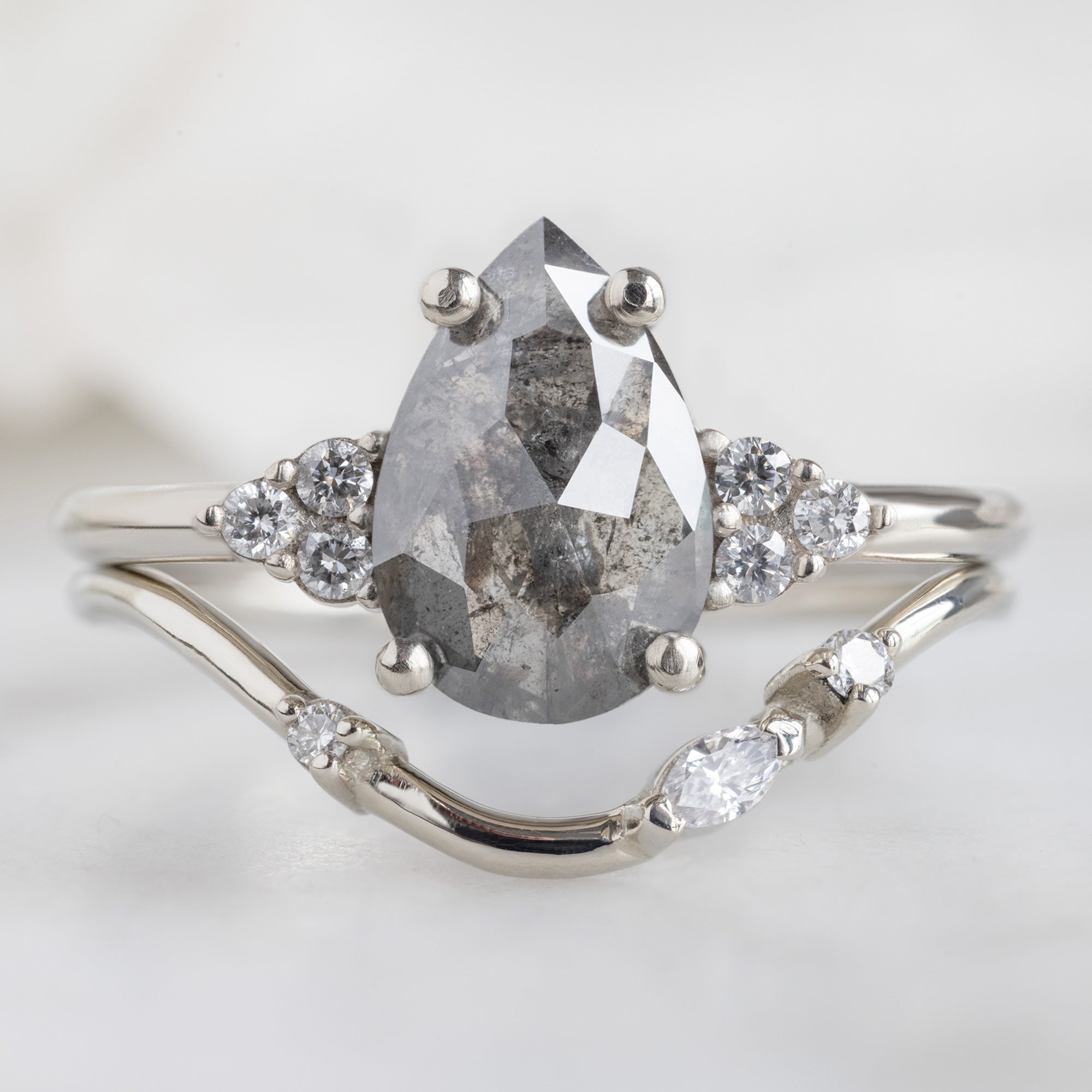 The Ivy Ring with a 1.46ct Pear Salt and Pepper Diamond in 14K White Gold Stacked with the Asymmetrical Marquise Diamond Arc Stacking Band on White Marble