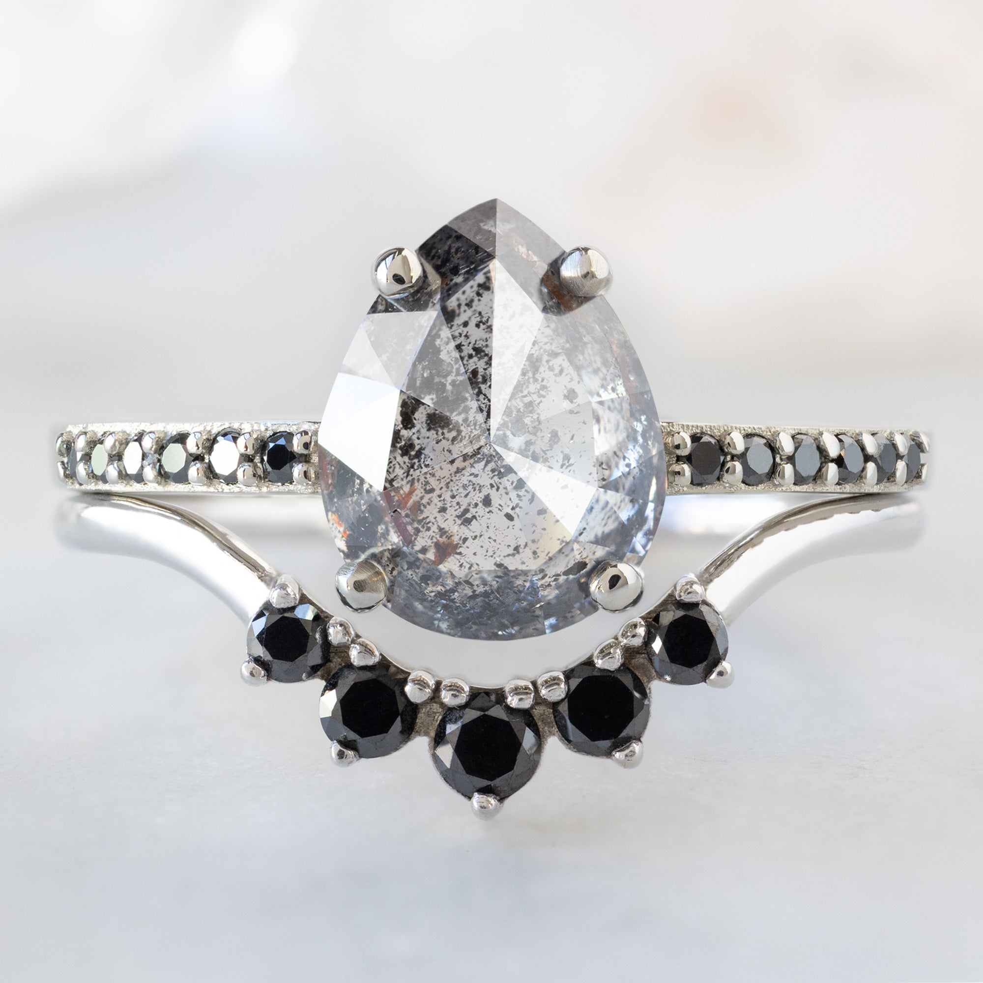 The Willow Ring with a 1.56ct Pear Salt and Pepper Diamond in 14K White Gold Stacked with the Round Black Diamond Sunburst Stacking Band on White Marble Surface