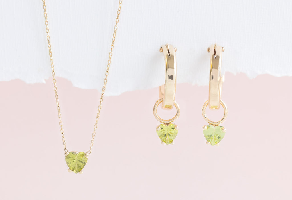 Yellow Gold Peridot Heart Hoops and Yellow Gold Sweetheart Peridot Necklace on White Textured Paper with Pink Background