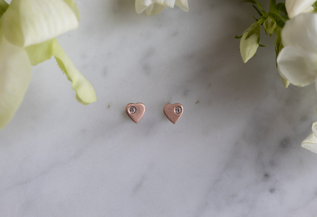 rose gold sweetheart diamond stud earrings on white marble tile with greenery surrounding 