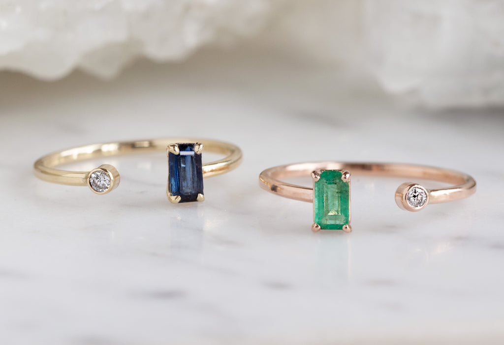 yellow gold open cuff sapphire and diamond ring and rose gold open cuff emerald and diamond ring on white marble tile